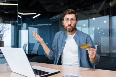 Portrait of disappointed and deceived businessman inside office, mature man looking at camera confused holding bank credit card and phone, employee got rejection error.