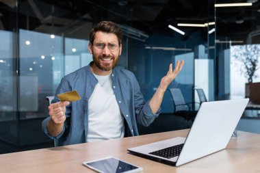 Portrait of disappointed and deceived businessman inside office, mature man looking at camera confused holding bank credit card and phone, employee got rejection error.
