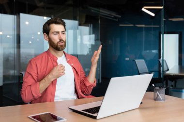 Its hot in the office, a businessman in a red shirt is hot, the air conditioner is not working, trying to cool down, a man inside the office is working with a laptop.