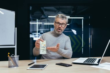 Portrait of mature businessman dissatisfied with income, gray haired man with dollar looking upset at camera, boss sad and disappointed working inside office with laptop