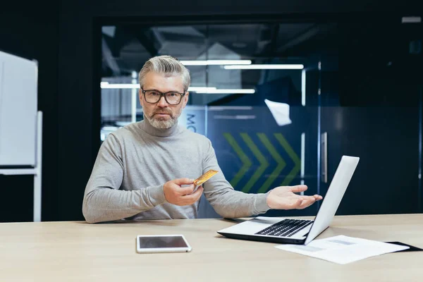 Portrait of disappointed man inside office, mature businessman received rejection and error of money transfer for online store purchase, boss looking confused at camera holding bank credit card.