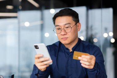 Serious and thinking asian man inside office using app on phone and bank credit card for online shop purchase and booking, businessman focused entering password at workplace.