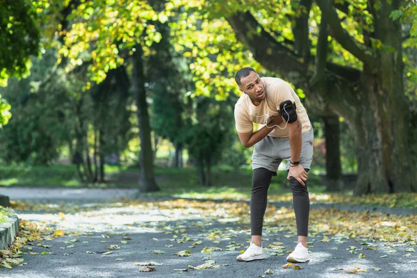 Sick man has severe heart pain after jogging, african american man holding hand to chest fast heart rate, tired sportsman on sunny day in park, using headphones and phone to listen to music.