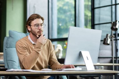 Portrait of a young designer, architect, engineer sitting in a modern office, working at a computer. He holds his chin with his hand, looks intently and thoughtfully at the monitor.