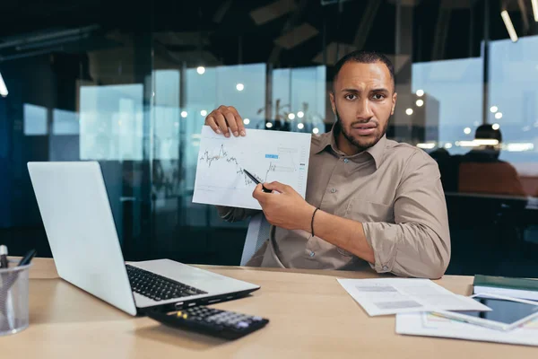 Portrait disappointed businessman inside office, african american man showing document graph with bad financial performance to camera, financier in overcoat working inside office using laptop.