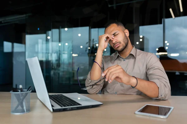 Tired and upset businessman inside office at work sitting at workplace with laptop, young african american financier took off glasses eyes hurt from overwork.