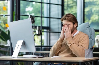 Tired office worker rubs eyes after long work at computer inside office, young blond businessman overworked at workplace in casual clothes.