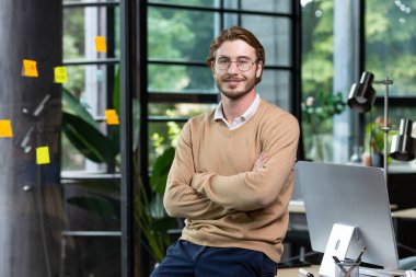 Portrait of successful smiling man inside office, blond man with crossed arms smiling and looking at camera at workplace with computer, businessman happy with achievement result.