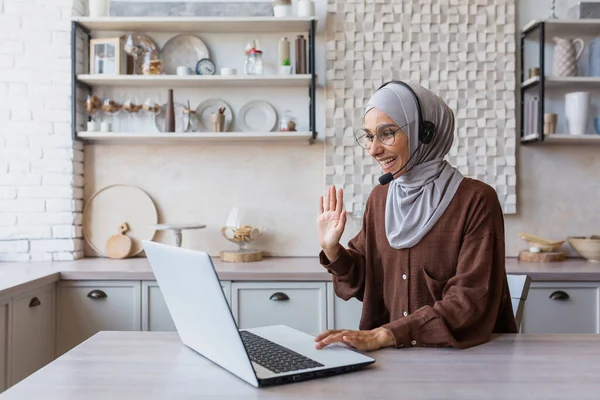 Woman in hijab working remotely from home sitting in kitchen using laptop and headset for video call, businesswoman smiling and talking to customers remotely customer support service online tech