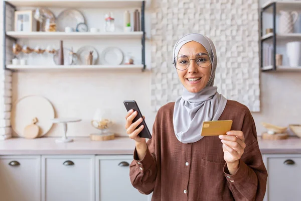 Portrait of happy online shopper woman at home, Muslim woman in hijab with phone smiling and looking at camera, Arab woman in kitchen holding bank credit card and phone.