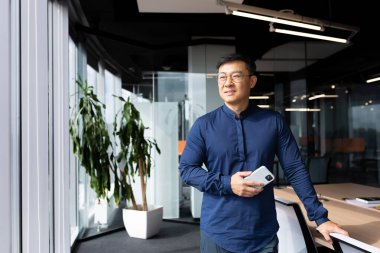 Asian man inside office standing near window dreaming and smiling, businessman in shirt and glasses thinking about future successful achievements and plans, boss at workplace holding phone.