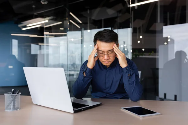 Stressed man working inside office, overworked asian man sitting at workplace sad and depressed holding hands on head, businessman frustrated thinking about problem.