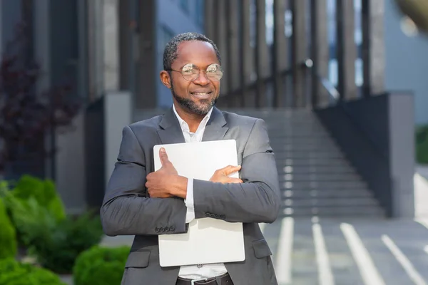 Portrait of African American male lawyer, legal advocate in suit and glasses standing posing outside near courthouse. Holds documents, papers with both hands. Smiling at the camera.