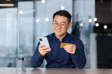 Satisfied and smiling man inside office at work, using app on phone and bank credit card for online shopping in online store, money transfer and booking, close up asian man with smartphone.