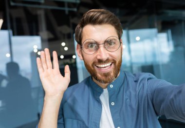 Mature man in office wearing shirt and glasses smiling and looking at smartphone camera, businessman taking selfie and talking on video call using smartphone, programmer waving hand greeting gesture.