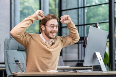 Smiling young male programmer, freelancer sits at the table, looks happy at the computer monitor. Raised his hands up, rejoices in success, victory.