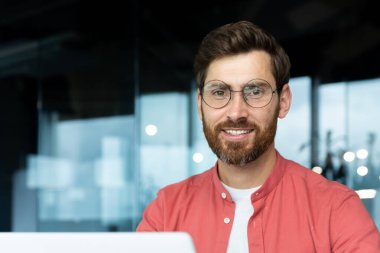 Close-up portrait of mature businessman inside office, man with beard and red shirt smiling and looking at camera, programmer developer coding software at workplace with laptop.