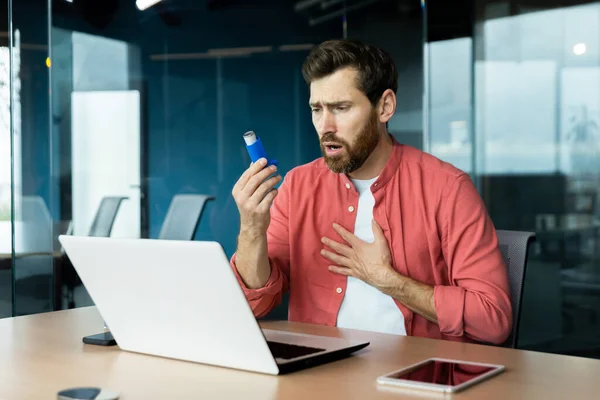 Asthma and respiratory problems in office worker, mature man coughing and using medicine in inhaler to ease breathing at workplace, mature businessman inside office with laptop.