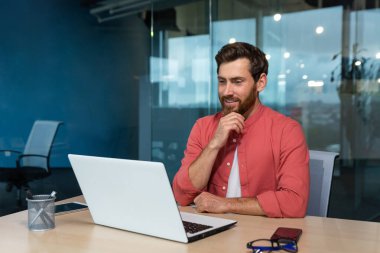 Portrait of a young male programmer, designer, freelancer in a red shirt who sits and works in the office at a laptop. He looks thoughtfully at the monitor, talks on a video call.