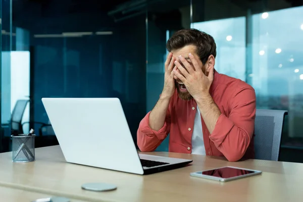 Problems at work. A young man sits in the office at a table with a laptop. Upset and tired, he holds his face with his hands. Screams with anger.