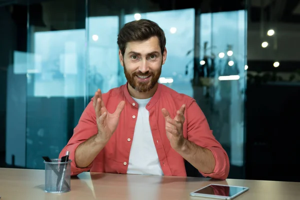 A young male teacher teaches online, conducts lectures, lessons. Sitting in the office at the table in front of the camera. Talks on a video call, explains, smiles.
