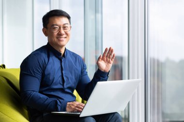 Portrait of successful smiling asian man inside office, man smiling and looking at camera near window, businessman working sitting with computer, waving hand in greeting gesture.