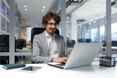 Cheerful and successful businessman inside office working with laptop, bearded man smiling and typing on laptop keyboard, programmer coding software in modern room.