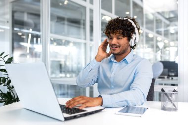 Young successful programmer inside modern office working with laptop, man in blue shirt smiling and happy, listening online music in headphones, audio books and podcasts, businessman satisfied work.