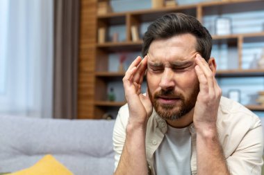 Headache of mature single man sitting on sofa alone at home and holding head with hands in living room.