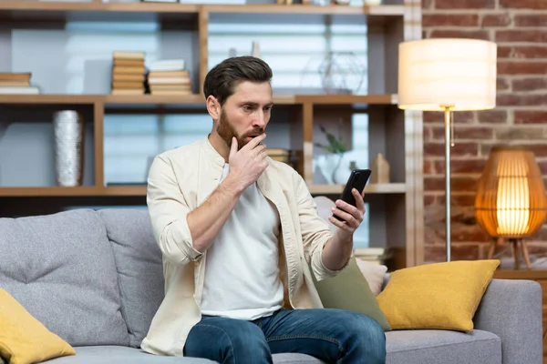 Sad and depressed man alone at home sitting on sofa, freelancer in casual clothes holding smartphone, unsatisfiedly reading bad news online from phone.