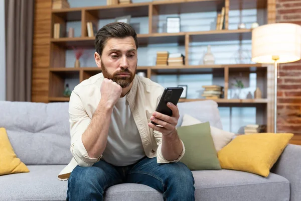 Sad and depressed man alone at home sitting on sofa, freelancer in casual clothes holding smartphone, unsatisfiedly reading bad news online from phone.