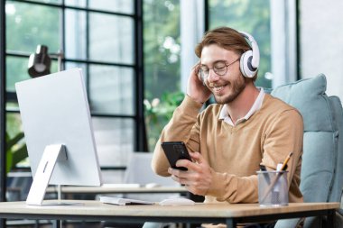 Smiling young male programmer, freelancer sits in the office at the table in headphones and holds the phone in his hand. Talks on a video call, listens to an audiobook, music, podcast.