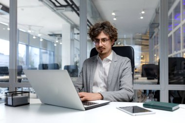 Serious and focused businessman inside modern office working on laptop, bearded man typing on keyboard, thinking man in business clothes sitting at workplace.