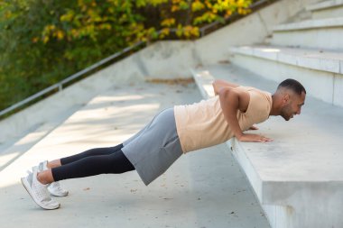 Cheerful and smiling hispanic man doing outdoor exercise in stadium, man stretching and recuperating before jogging in summer park.