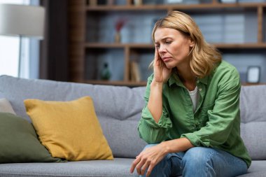 Mature woman alone at home with boss and depressed, housewife sitting upset on sofa holding head with hands in living room.