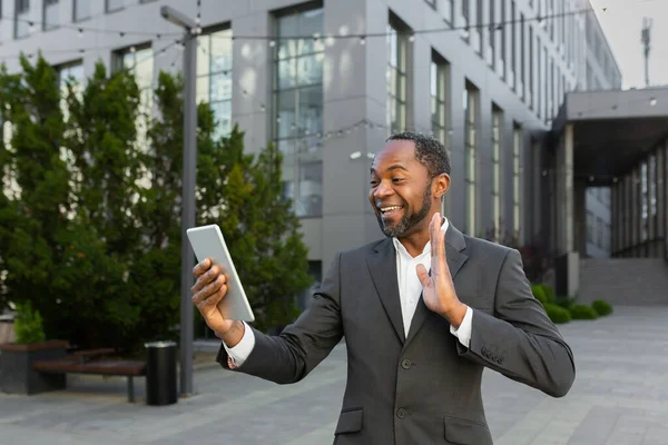 An African-American businessman is standing outside in a suit, holding a tablet and talking on a video call.