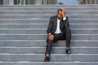 Sad african american businessman sitting on stairs outside office building, man in business suit bankrupt fired from job, boss lost money.