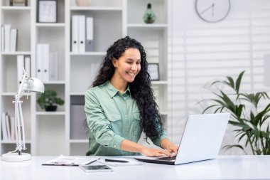 Young successful hispanic businesswoman working in bright home office indoors sitting at table with documents and reports, woman using laptop at work happy smiling.