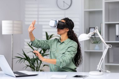 Young successful woman inside the office using vr glasses, virtual reality simulator cross charts and documents online, latin american woman using laptop at work.