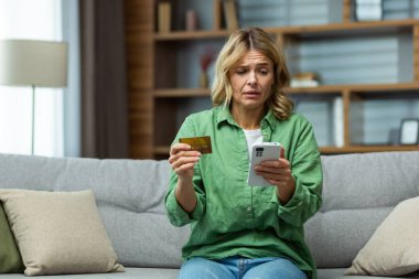 A woman sitting on sofa at home. Worriedly holding phone and credit card. Problem with the account, online payment, blocking, bankruptcy, fraud.