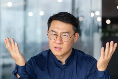 Angry boss close-up talking on video call using laptop, man looking at web camera and arguing nervous businessman unhappy with online meeting with colleagues, asian man at work inside office angry.