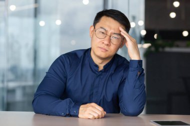Portrait of overworked and tired office worker at workplace, asian man in glasses and shirt looking upset at camera, businessman not satisfied with work and own results achievement in depression.