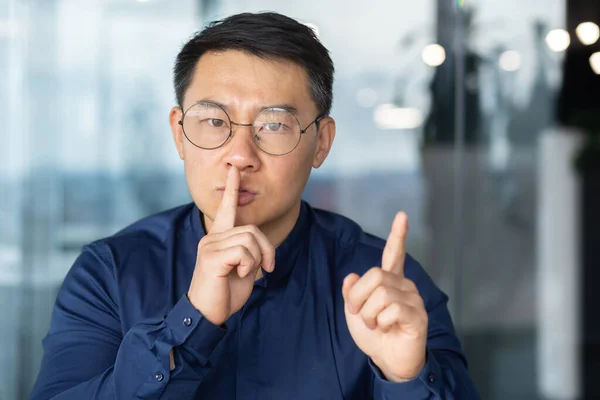 Close up portrait of man holding finger near mouth demanding silence at workplace, Asian man inside office looking at camera upset and worried trying to concentrate.