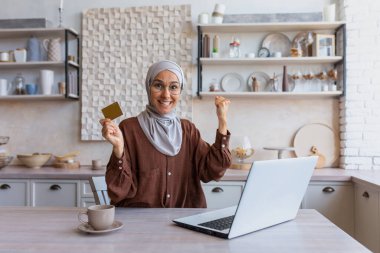 Happy young beautiful Muslim woman in hijab sitting at laptop at home at table, holding and showing credit card in hand, smiling at camera, showing yes hand gesture.