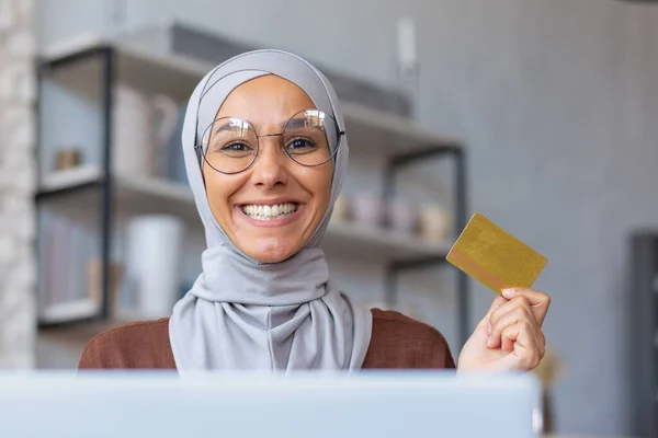 Portrait of a young beautiful Muslim woman in a hijab sitting at a laptop, holding and showing a credit card in her hand, smiling at the camera.