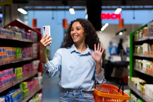 Shopping. A young Latin American woman walks in a supermarket between rows of shelves with a shopping basket. Talks on the phone, video call,