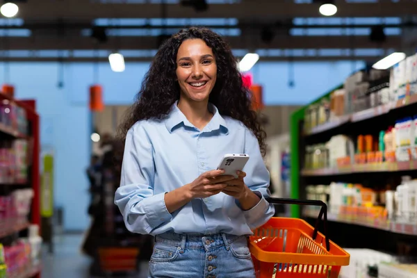 Shopping in a supermarket. A young Latin American woman stands between rows of shelves with a cart. He holds the phone, has a list, writes messages, orders and pays online. Smiling at the camera.
