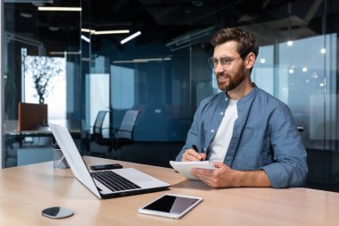 Portrait of a young businessman working in the office using a laptop, writing down information in a notebook, talking on a video call.