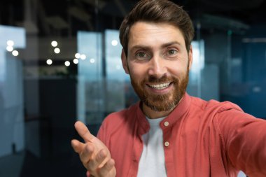 Close-up photo. Young businessman, freelancer, blogger in a red shirt in the office talking on a video call from the phone, smiling at the camera.