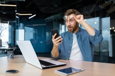 Genç bir adam masadaki bir ofiste dizüstü bilgisayar kullanarak çalışıyor. Telefonu elinde tutuyor, mutlu ve şok olmuş bir şekilde ekrana bakıyor, gözlüğünü şaşkınlıkla yüzüne kaldırıyor. İyi haberlerim var..